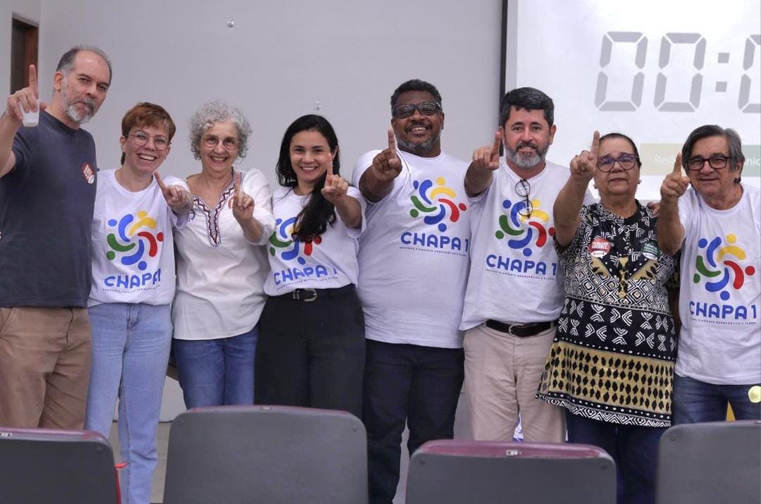 Esta é uma foto com um grupo de oito pessoas - quatro homens e quatro mulheres - alinhadas lado a lado, todas sorrindo e fazendo o gesto do número 1 com o dedo indicador levantado. Quatro delas estão usando camisetas brancas com o logotipo colorido e o texto CHAPA 1 estampados, enquanto as outras estão vestidas com roupas casuais ou tradicionais. A imagem parece ter sido tirada em um ambiente interno, com cadeiras visíveis na parte inferior e um projetor ou tela ao fundo, onde aparece um número digital 00:00. O clima é descontraído e de celebração, possivelmente relacionado a uma campanha ou evento coletivo.