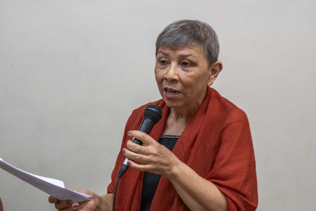 Foto da profesora Graciela Chamorro, mulher idosa, parda, de cabelos grisalhos e cortados bem curtos. Fotografada acima do abdômen em frente a uma parede branca, ela usa um casaco cor de tijolo sobre uma blusa preta. Ela segura um microfone com a mão esquerda e folhas de papel branco com a mão direita.