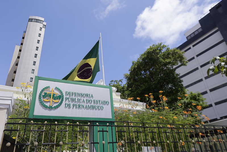 A imagem mostra a fachada da Defensoria Pública do Estado de Pernambuco, uma instituição que oferece assistência jurídica gratuita. Em destaque, há uma placa verde e branca com o nome da instituição e o símbolo da balança da justiça cercada por folhas verdes. Ao fundo, vê-se um prédio alto e claro ao lado de um edifício mais escuro, além de uma grande árvore com flores alaranjadas. Uma bandeira do Brasil tremula acima da placa, destacando-se pelo verde, azul e branco. O céu é azul com poucas nuvens, indicando um dia ensolarado. A foto foi tirada de um ângulo baixo, enfatizando a placa e a bandeira.