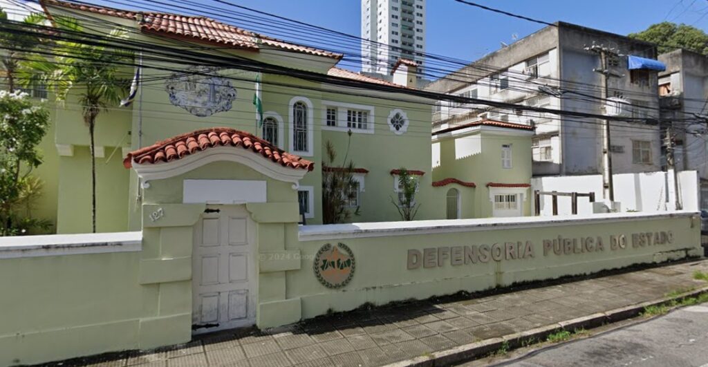 Essa imagem mostra a fachada de um prédio público com arquitetura clássica, pintado na cor verde claro e com detalhes em branco. Há uma porta central, em arco, com telhado de telhas vermelhas em estilo colonial. Na fachada do muro está escrito DEFENSORIA PÚBLICA DO ESTADO, indicando que se trata de um prédio vinculado à Defensoria Pública, instituição responsável por prestar assistência jurídica gratuita à população que não pode pagar por serviços advocatícios. No muro, à esquerda, há um símbolo representando a instituição e o número 127 indicando o endereço do local. A imagem também mostra fios elétricos cruzando a cena, árvores ao redor do prédio e, ao fundo, é possível ver edifícios residenciais e comerciais modernos. O céu está limpo e azul, sugerindo um dia ensolarado.