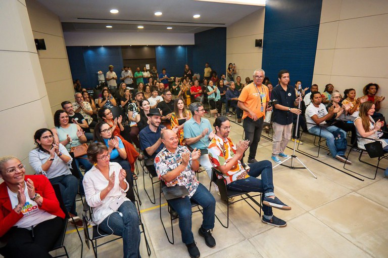 A imagem mostra uma sala de conferências com muitas pessoas sentadas em cadeiras, organizadas em fileiras. A maioria parece estar aplaudindo algo ou alguém. As pessoas têm diferentes idades e estilos, e algumas estão sorrindo. No fundo, há uma parede azul e um grupo menor de pessoas de pé, assistindo à cena. Há também um homem em pé, perto de um microfone em um pedestal. O ambiente é bem iluminado, com luzes no teto.