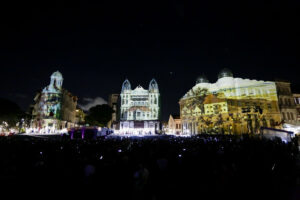 A foto captura um evento noturno em um espaço público cercado por edifícios históricos. As fachadas dos prédios estão sendo utilizadas como telas para projeções mapeadas, exibindo imagens de cenas históricas, paisagens e possivelmente pinturas relacionadas a contextos culturais ou coloniais. No centro, um edifício com arquitetura marcante tem torres simétricas e está completamente iluminado por uma projeção. À esquerda e à direita, outros prédios também exibem imagens coloridas, criando um efeito visual impressionante. Uma grande multidão está presente, assistindo ao espetáculo, com algumas pessoas registrando o momento com celulares. A iluminação e o uso das projeções contrastam com o céu escuro da noite, enfatizando a importância das imagens e da arquitetura no evento.