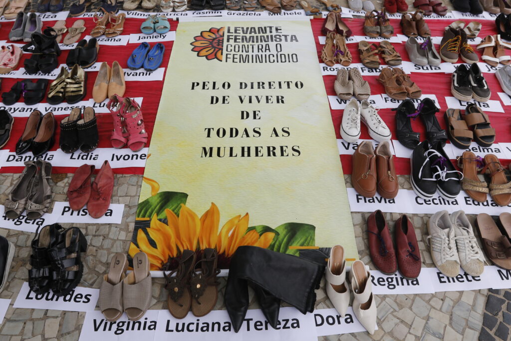 A imagem mostra uma instalação com sapatos femininos dispostos no chão, cada par acompanhado de um nome feminino em papel. Ao centro, há um cartaz com a frase: Levante Feminista Contra o Feminicídio - Pelo direito de viver de todas as mulheres, decorado com um girassol. A instalação simboliza as vítimas de feminicídio, promovendo conscientização e justiça. O chão é de pedra, e os sapatos variam em estilo e cores, representando a diversidade das mulheres homenageadas.