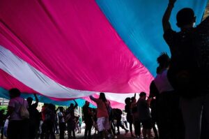 Esta é uma foto tirada de baixo de uma grande bandeira com as cores da bandeira do orgulho trans (azul claro, rosa e branco). A perspectiva mostra várias pessoas caminhando ou paradas sob a bandeira, levantando-a com as mãos, criando uma sensação de união e celebração. As silhuetas das pessoas aparecem contra a luz filtrada pela bandeira, sugerindo que o evento acontece ao ar livre, possivelmente em um protesto ou marcha.