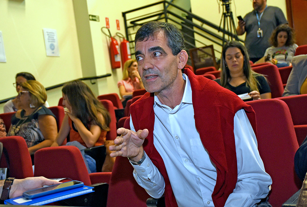 A imagem mostra Adilson Paes de Souza, homem branco, de meia idade, em destaque, sentado em uma sala de palestras ou auditório com cadeiras vermelhas. Ele está falando e gesticulando com uma expressão engajada, enquanto usa uma camisa branca e tem um casaco vermelho jogado sobre os ombros. Atrás dele, outras pessoas estão sentadas, algumas aparentam estar prestando atenção ou interagindo com algo, como escrevendo ou olhando em suas mãos. No fundo, há uma escada, extintores de incêndio fixados na parede e um ambiente iluminado por luz artificial, sugerindo que a foto foi tirada em um evento ou reunião em um espaço fechado.