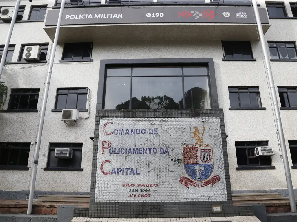 A imagem mostra a fachada de um prédio institucional com o nome Polícia Militar destacado na parte superior. No centro, há uma entrada ampla com janelas de vidro, acima da qual está o brasão da Polícia Militar de São Paulo. Em frente à entrada, há um mosaico com os dizeres Comando de Policiamento da Capital (CPC), acompanhado de um brasão colorido e uma inscrição que menciona São Paulo, Jan 2004, 450 anos, referindo-se ao aniversário da cidade. O prédio é revestido com paredes claras, possui janelas de moldura preta, e há mastros para bandeiras nas laterais.