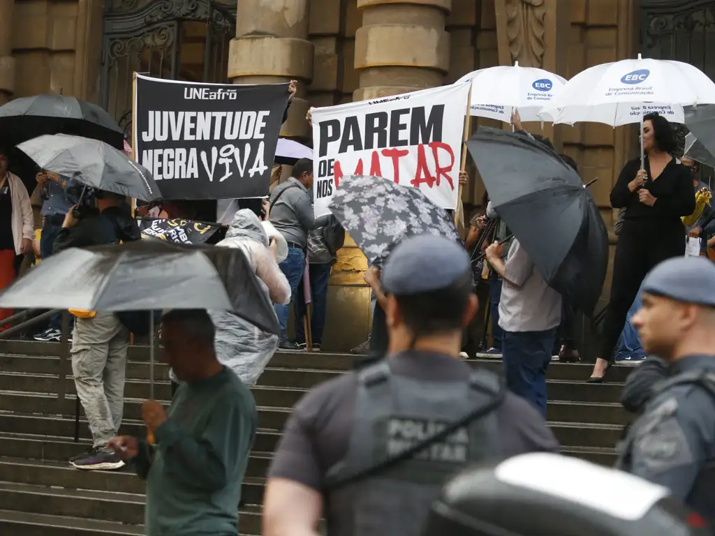 Na imagem, um grupo de pessoas está reunido em frente a um prédio de arquitetura clássica, com escadarias amplas. Algumas pessoas seguram cartazes e faixas, enquanto outras estão com guarda-chuvas abertos, indicando que está chovendo. Entre os cartazes, destacam-se os dizeres: JUVENTUDE NEGRA VIVA e PAREM DE NOS MATAR. Há também a presença de policiais militares niformizados em primeiro plano, observando a manifestação. A cena transmite a ideia de um protesto pacífico, possivelmente contra a violência policial ou questões de racismo e injustiça social.