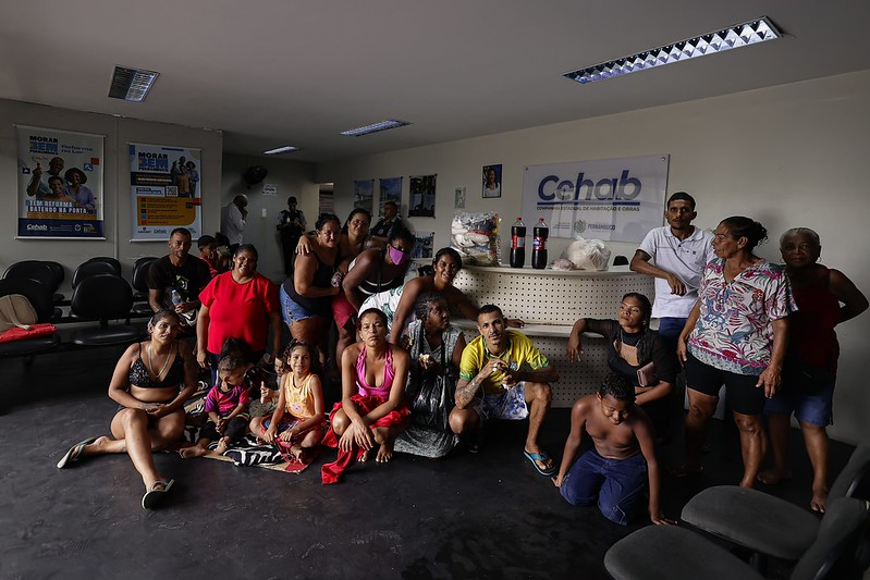 A imagem mostra um grupo de pessoas, incluindo mulheres, crianças e homens, posando juntas em frente ao balcão da sala de espera da Cehab.