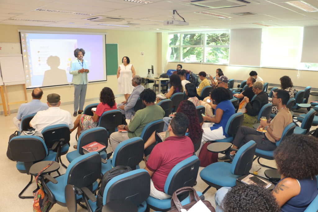 A imagem mostra uma sala de aula com várias pessoas sentadas em cadeiras azuis, voltadas para a frente da sala. Na frente, duas pessoas estão de pé, aparentemente apresentando algo. Há um projetor ligado, exibindo uma apresentação na tela. A sala é bem iluminada, com luz natural entrando pelas janelas à direita. As pessoas na sala parecem estar prestando atenção à apresentação.