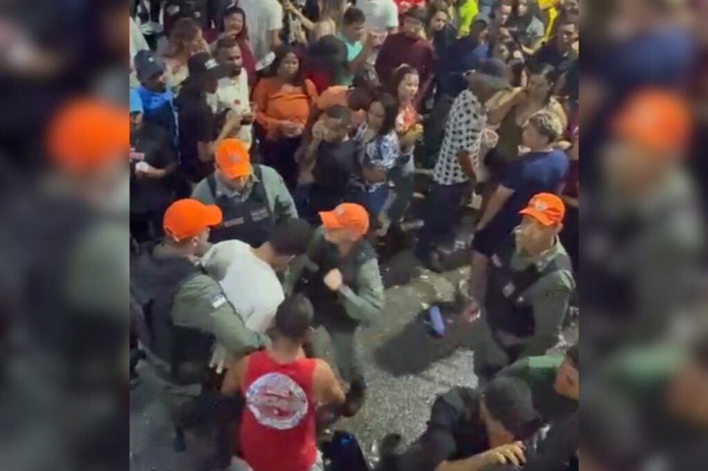 A imagem mostra uma cena em meio a uma grande aglomeração de pessoas em um evento ou festa de rua. No centro da imagem, há vários agentes de segurança usando uniformes verdes e bonés laranjas, aparentemente tentando conter um homem de camiseta branca. O homem parece estar sendo segurado por pelo menos dois agentes, enquanto um terceiro parece estar pronto para agir. Há muitas pessoas ao redor assistindo à situação, algumas com expressões de surpresa ou preocupação. A iluminação e a movimentação indicam que o evento ocorre à noite.