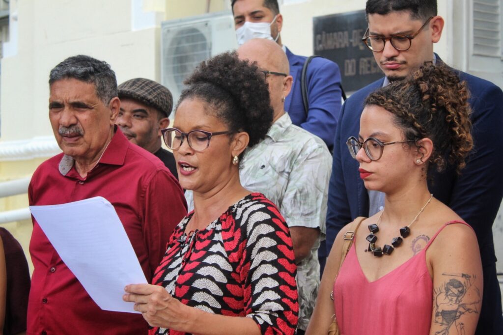 Uma mulher de cabelo crespo preso e óculos lê um documento em voz alta, acompanhada por outras pessoas ao fundo. Ela veste uma blusa estampada em vermelho, branco e preto. Ao lado dela, está um homem de camisa vermelha e outra mulher de vestido vermelho com tatuagens no braço. O grupo parece estar participando de um evento público em frente a um prédio oficial.