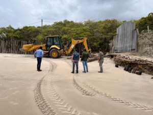 A imagem mostra uma praia com marcas de pneus na areia. No centro, há uma retroescavadeira amarela parada, com quatro homens observando a cena. Eles estão de pé, posicionados de costas ou de lado para a câmera, olhando em direção à retroescavadeira e a uma estrutura de madeira e pedra na lateral direita. À esquerda, há uma cerca feita de troncos de madeira, e ao fundo, é possível ver vegetação densa. O céu está nublado, dando um aspecto cinzento ao ambiente.