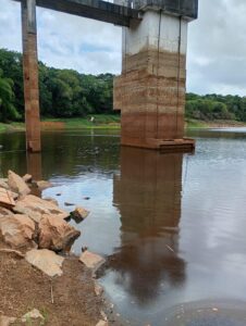 Esta foto mostra uma cena ao ar livre em um ambiente natural com uma estrutura de concreto parcialmente submersa em água. A estrutura parece ser uma ponte ou pilar, com marcas visíveis de diferentes níveis de água ao longo de sua superfície, indicando variações no nível do rio ao longo do tempo. À esquerda, há pedras irregulares na margem do rio, e ao fundo, uma vegetação densa e verde cobre o restante do cenário. O céu parece nublado, sugerindo um clima úmido ou recente chuva. A água é calma e reflete parte da estrutura de concreto e da vegetação ao redor.