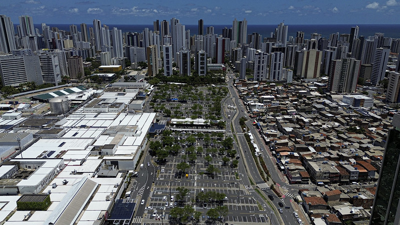 A imagem aérea de Boa Viagem, no Recife, mostra um forte contraste urbano: de um lado, prédios modernos e um grande shopping; do outro, uma comunidade de moradias simples. O cenário reflete desigualdade socioeconômica e evidencia o fenômeno da ilha de calor, com áreas verticalizadas e pavimentadas retendo mais calor do que as regiões arborizadas. Ao fundo, o oceano destaca a localização litorânea da cidade.