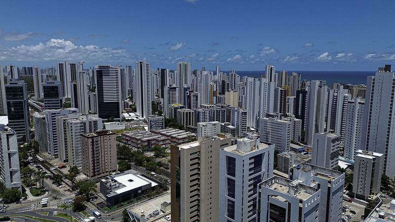 A imagem aérea mostra uma área densamente urbanizada com muitos prédios altos e modernos, predominantemente brancos e cinzas. O mar azul aparece no horizonte, contrastando com a paisagem urbana. O céu está limpo e a forte iluminação realça sombras e reflexos nos edifícios. A cena destaca o adensamento urbano e o fenômeno da ilha de calor, causado pela falta de vegetação e o excesso de concreto e asfalto.