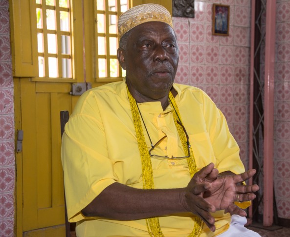 Foto de Ivo de Xambá. Ele é um homem negro e idoso vestindo roupas amarelas e um colar dourado. Ele usa um chapéu tradicional e está sentado em um ambiente interno, com paredes de azulejos decorados e uma porta amarela ao fundo. Ele parece estar em uma conversa ou entrevista, com as mãos gesticulando.