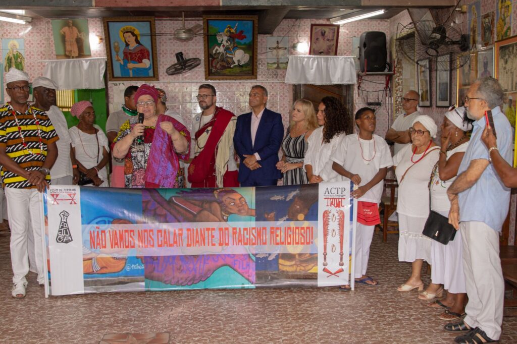 A imagem mostra um grupo de pessoas reunidas em um ambiente interno, possivelmente um centro religioso ou cultural. Algumas estão vestidas com roupas tradicionais brancas e outras com roupas coloridas. No centro, há um banner com a frase NÃO VAMOS NOS CALAR DIANTE DO RACISMO RELIGIOSO! em letras vermelhas, acompanhado por imagens e símbolos religiosos. A cena parece ser um evento ou manifestação contra o racismo religioso, destacando a importância da luta contra a discriminação baseada na religiã