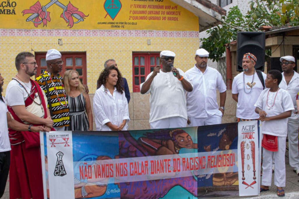 A imagem mostra um grupo de pessoas reunidas em frente a um prédio decorado com elementos culturais e tradicionais. Há um banner com a frase “NÃO VAMOS NOS CALAR DIANTE DO RACISMO RELIGIOSO!” e imagens que destacam a temática da luta contra a intolerância religiosa. Algumas pessoas usam trajes brancos típicos de religiões de matriz africana, enquanto outras estão em roupas casuais ou estampadas. O evento é uma manifestação em apoio à diversidade religiosa e contra o racismo.