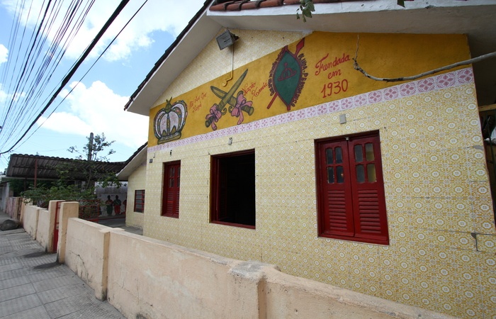 A imagem mostra uma construção simples com fachada decorada por azulejos amarelos e um painel pintado com símbolos e a inscrição Fundada em 1930. As janelas são de madeira vermelha e há um muro baixo na frente, em um ambiente residencial tranquilo.