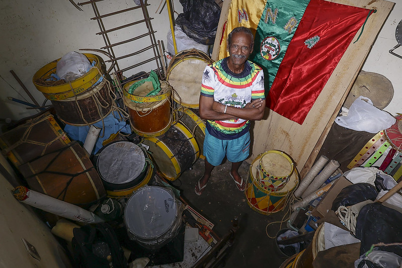 A foto mostra mestre Walter de França, homem negro idoso de cabelos grisalhos, vestindo uma camiseta colorida e calção azul, de braços cruzados e expressão serena. Ele está descalço em um pequeno cômodo repleto de tambores e instrumentos de percussão, muitos com detalhes amarelos e pretos. Ao fundo, há uma grande bandeira verde e vermelha com bordados. O espaço parece um depósito de instrumentos musicais, com objetos empilhados e materiais espalhados.