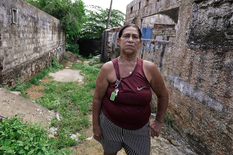 A imagem mostra Ednalva Lopes, uma mulher de aparência séria, pele clara e cabelos escuros presos para trás. Ela veste uma blusa regata vermelha e shorts listrados em preto e branco, com um molho de chaves pendurado no ombro. O cenário ao redor é um ambiente degradado, com paredes de tijolos gastos e sem acabamento, além de um chão irregular com mato crescendo. Ao fundo, há uma passagem estreita entre os muros, levando a uma área com mais vegetação.