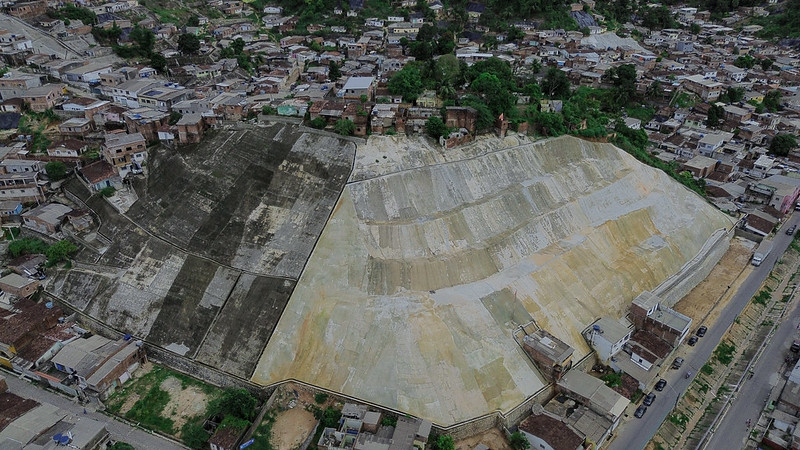 A imagem mostra uma vista aérea de uma área urbana com muitas casas e construções. No centro da imagem, há uma grande área dividida em duas seções distintas. A seção à esquerda é mais escura e parece ser uma encosta ou colina com vegetação esparsa e algumas áreas de concreto. A seção à direita é mais clara e parece ser uma área de concreto ou pavimentada, possivelmente uma obra de contenção ou estabilização de solo. Ao redor dessas duas seções, há muitas casas e ruas, indicando que se trata de uma área densamente povoada.