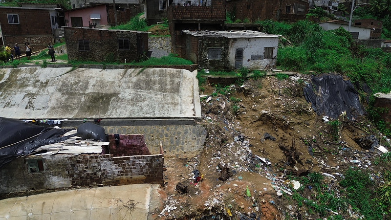 A imagem mostra uma área residencial em uma encosta, onde há sinais de deslizamento de terra. Na parte inferior da imagem, há casas danificadas, uma delas com o telhado parcialmente destruído e coberto por uma lona preta. A encosta está coberta por detritos e lixo, indicando um deslizamento recente. No topo da encosta, há mais casas, algumas em condições precárias. A vegetação ao redor é densa, e há pessoas ao fundo, possivelmente avaliando os danos.