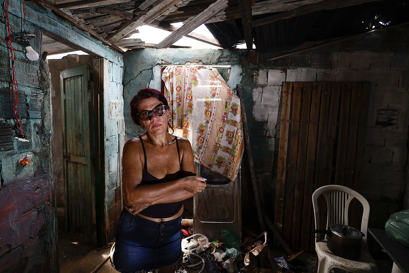 A imagem mostra Maria José, uma mulher de meia-idade, com pele clara, cabelos avermelhados e óculos escuros. Ela veste uma blusa preta de alças finas e uma saia ou short jeans escuro, com uma expressão séria e braços cruzados, segurando algo na mão. O ambiente é uma casa em condições precárias, com um teto parcialmente destruído, paredes de tijolos aparentes e objetos espalhados pelo chão.