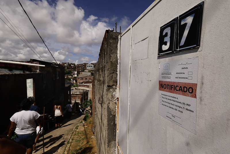 A imagem mostra uma rua estreita e inclinada em uma área urbana, com várias casas ao longo do caminho. Do lado direito da imagem, há um muro branco com o número 37 em preto e uma placa com a palavra NOTIFICADO em destaque. A placa parece ser um aviso oficial. Algumas pessoas estão caminhando pela rua, descendo a ladeira. O céu está azul com algumas nuvens brancas e, ao fundo, é possível ver mais casas e construções.