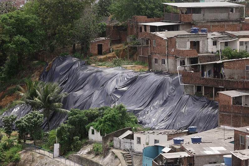 A imagem mostra uma área residencial em uma encosta. Várias casas de tijolos aparentes estão construídas na encosta, que está coberta com uma grande lona preta, provavelmente para proteger o solo de deslizamentos ou erosão. Há vegetação ao redor, incluindo árvores, arbustos e algumas palmeiras em primeiro plano. As casas variam em tamanho e estrutura, algumas com telhados de zinco e outras com telhados de concreto.