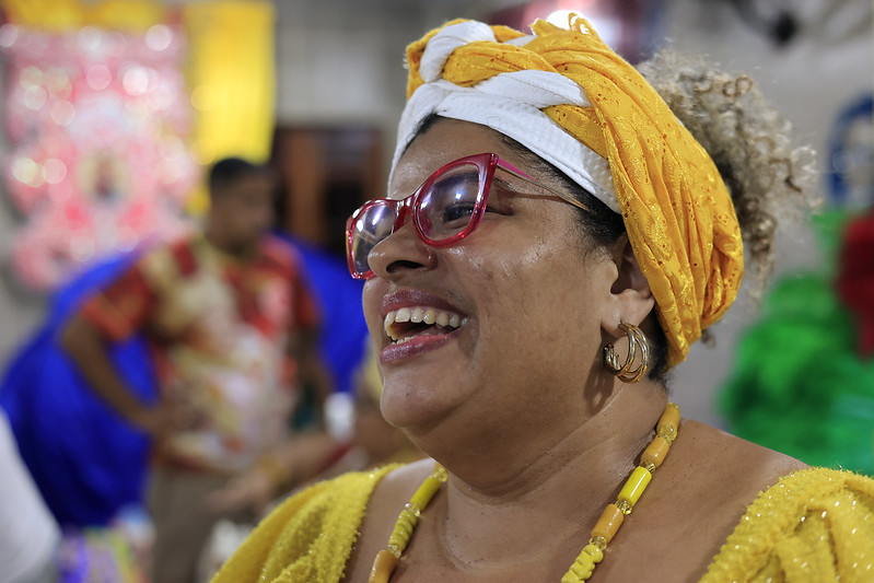 A imagem mostra uma mulher negra vestida com um turbante amarelo e branco, brincos grandes, um colar de contas amarelas e uma roupa amarela brilhante. Atrás dela, há outra pessoa com uma camisa colorida e um ambiente decorado com cores vibrantes.