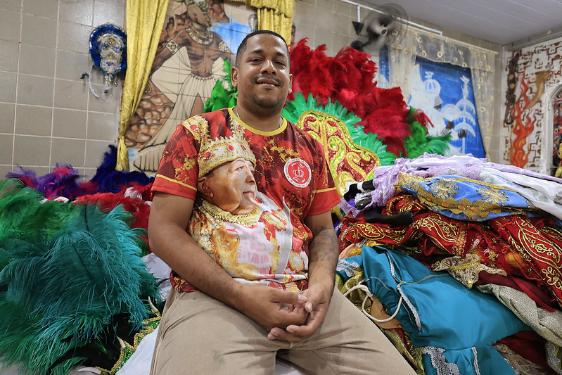 Um homem jovem, de pele negra, está sentado em meio a roupas e adereços coloridos de carnaval. Ele veste uma camisa vermelha com a imagem de um rei e sorri levemente. Ao fundo, há painéis decorados com temas africanos e religiosos.