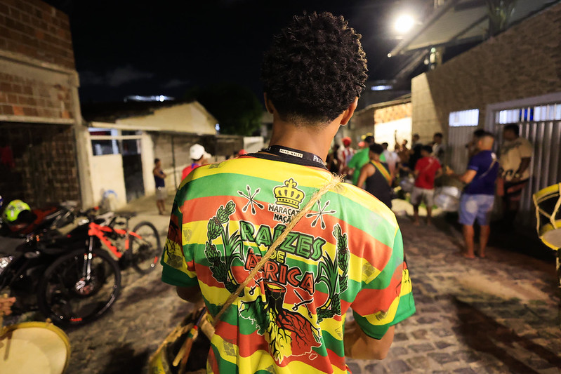 Jovem negro de costas com uma camisa verde, vermelha e amarela, com a frase Maracatu Nação Raízes de África. Ele está em um ambiente aberto, numa rua de paralelepípedos, segurando um instrumento de percussão.