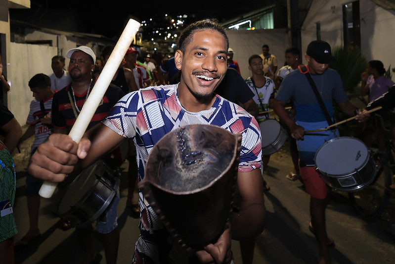 Jovem negro sorrindo segurando um gonguê, instrumento musical que marca o maracatu. Ele veste uma camisa azul, rosa e branca e está em um ensaio aberto.