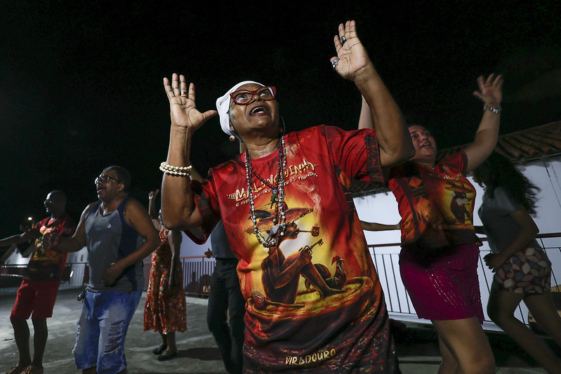 A foto mostra uma mulher idosa dançando com energia em uma festa noturna ao ar livre. Ela usa óculos vermelhos, um lenço branco na cabeça e uma camisa vermelha estampada. Ela está olhando para cima e com as mãos para o alto. Seu gestual expressivo transmite alegria e confiança. Ao fundo, outras pessoas também dançam, vestindo roupas coloridas. O evento parece ser uma celebração cultural ou religiosa, possivelmente afro-brasileira. A iluminação destaca as cores vibrantes e a animação do momento.