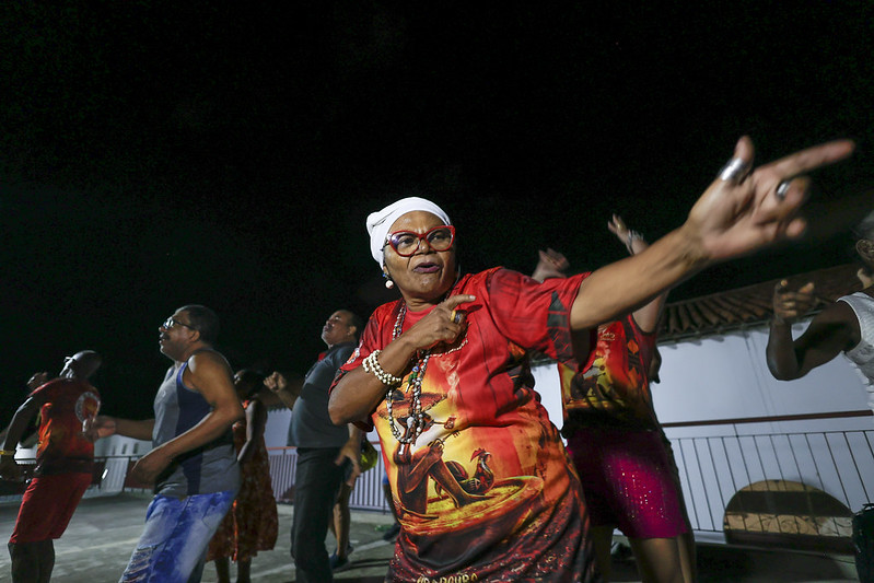 A foto mostra uma mulher idosa dançando com energia em uma festa noturna ao ar livre. Ela usa óculos vermelhos, um lenço branco na cabeça e uma camisa vermelha estampada. Seu gestual expressivo transmite alegria e confiança. Ao fundo, outras pessoas também dançam, vestindo roupas coloridas. O evento parece ser uma celebração cultural ou religiosa, possivelmente afro-brasileira. A iluminação destaca as cores vibrantes e a animação do momento.