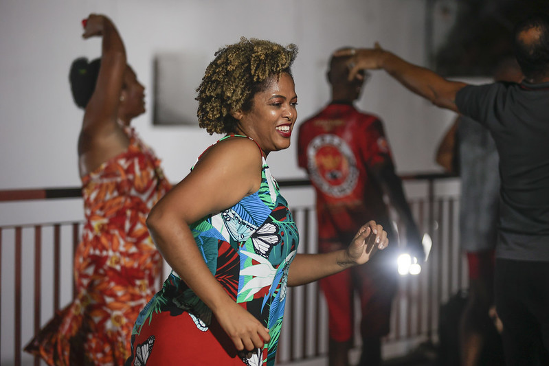 A foto mostra uma mulher jovem e sorridente dançando com entusiasmo em uma festa ao ar livre à noite. Ela tem cabelo cacheado e veste uma blusa azul com estampas tropicais. Seu corpo está em movimento, transmitindo alegria. Ao fundo, outras pessoas também dançam, incluindo uma mulher de vestido vermelho estampado e um homem de camiseta vermelha. Os gestos dos participantes sugerem uma celebração cultural animada. A iluminação destaca os rostos e roupas,