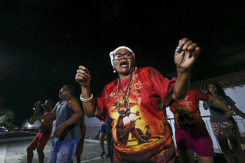 A foto mostra uma mulher idosa dançando com energia em uma festa noturna ao ar livre. Ela usa óculos vermelhos, um lenço branco na cabeça e uma camisa vermelha estampada. Seu gestual expressivo transmite alegria e confiança. Ao fundo, outras pessoas também dançam, vestindo roupas coloridas. O evento parece ser uma celebração cultural ou religiosa, possivelmente afro-brasileira. A iluminação destaca as cores vibrantes e a animação do momento.