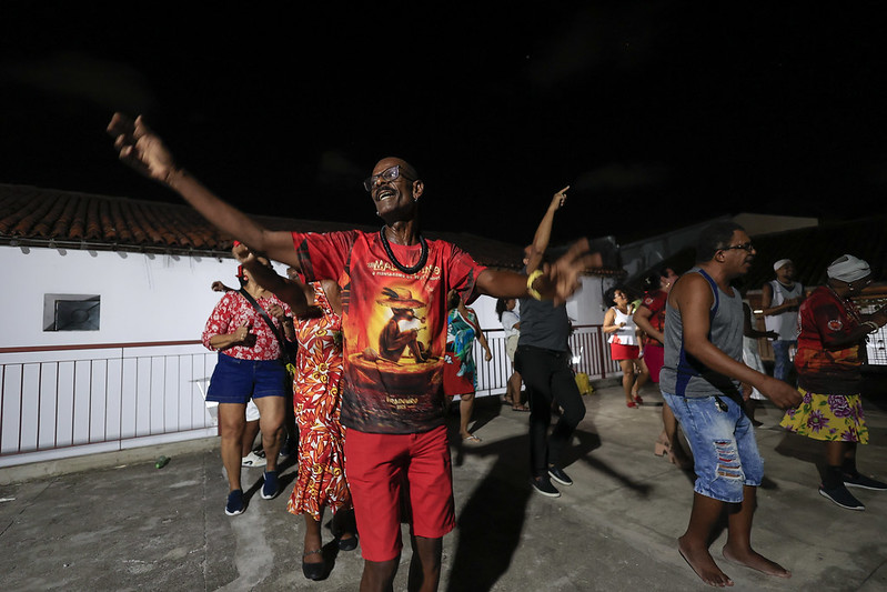 A foto mostra um homem alto, magro e sorridente dançando com os braços abertos em uma festa ao ar livre à noite. Ele usa óculos, uma camisa vermelha estampada com uma imagem marcante e bermuda vermelha. Ao fundo, várias pessoas de diferentes idades também dançam animadamente, vestindo roupas coloridas. Algumas usam roupas estampadas, enquanto outras estão de shorts e camisetas casuais. A festa acontece em um espaço aberto com um muro branco e telhados ao redor.