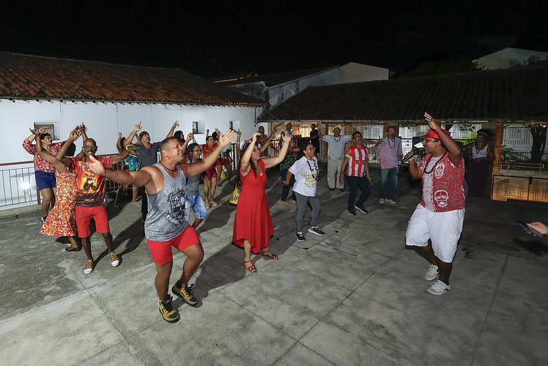 A imagem mostra um grupo de pessoas dançando em um espaço ao ar livre durante a noite. O local parece ser um pátio com piso de concreto e telhados de telha. As pessoas estão vestidas de maneira casual e colorida, com algumas usando roupas vermelhas e outras com roupas variadas. Elas estão com os braços levantados e parecem estar seguindo uma coreografia ou participando de uma atividade de dança em grupo. Ao fundo, há uma construção com paredes brancas e telhado de telha, e algumas pessoas estão observando a dança. A iluminação é suficiente para ver claramente as pessoas e o ambiente ao redor.