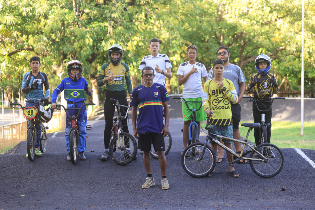 A foto mostra um grupo de 9 pessoas em uma pista de BMX, cercada por árvores verdes. Seis delas estão com bicicletas e roupas esportivas, incluindo capacetes e camisetas estampadas, algumas com a bandeira do Brasil. Dois homens sem bicicletas estão ao fundo, vestindo roupas casuais. Todos estão posando para a foto, alguns fazendo gestos com as mãos. O solo da pista é de terra batida ou asfalto escuro. O clima parece ensolarado, com sombras das árvores ao fundo.