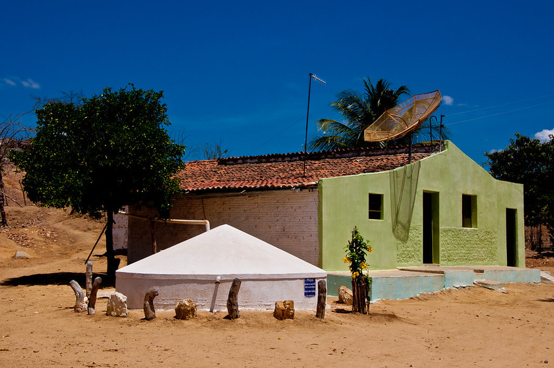 A imagem mostra uma casa simples em uma área rural. A casa tem paredes brancas e uma fachada pintada de verde claro. O telhado é de telhas vermelhas e há uma antena parabólica instalada no telhado. Em frente à casa, há a estrutura branca com um telhado em forma de pirâmide da cisterna para captar água de chuva. Ao lado dessa estrutura, há uma árvore com folhas verdes. O chão ao redor é de terra seca e há algumas pedras e troncos de madeira espalhados. O céu está claro e azul.