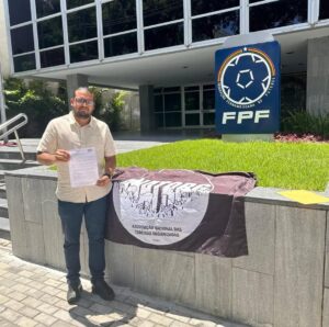 Foto de Adriano Costa em pé, segurando um documento, em frente a um prédio moderno com fachada de vidro. Ele é um homem pardo, de cabelos escuros e barba rala, usando óculos. Ao lado dele há uma bandeira estendida sobre um parapeito de concreto. A bandeira tem o logotipo da Associação Nacional das Torcidas Organizadas (ANATORG) e a palavra BRASIL escrita abaixo. À direita da pessoa, há uma placa grande com o logotipo da Federação Pernambucana de Futebol (FPF). A pessoa está vestida com uma camisa de manga curta bege e calça jeans. A imagem parece ter sido tirada em um dia ensolarado.