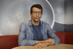 Foto de Humberto Barbosa, homem branco, de cabelos pretos lisos e curtos, usando óculos de aros escuros. Ele está sentado com as mãos postas sobre uma mesa de tampo de madeira, usando casaco jeans e camisa azul marinho.