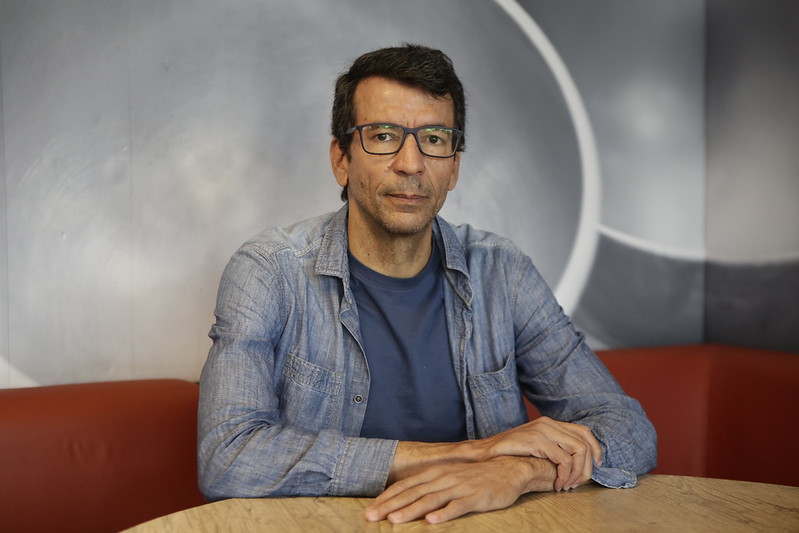 Foto de Humberto Barbosa, homem branco, de cabelos pretos lisos e curtos, usando óculos de aros escuros. Ele está sentado com as mãos postas sobre uma mesa de tampo de madeira, usando casaco jeans e camisa azul marinho.