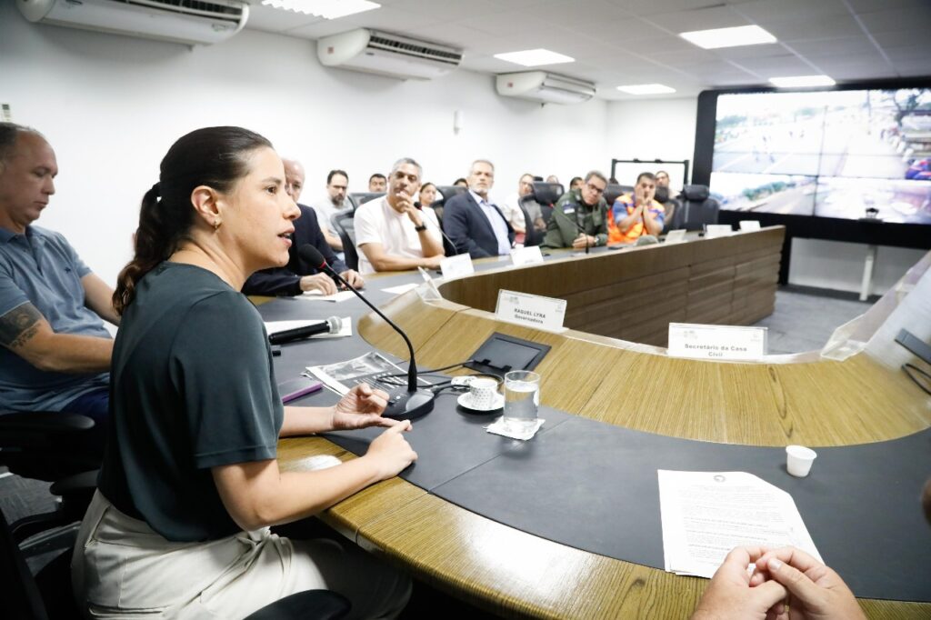 A imagem mostra a governadora Raquel Lyra em uma reunião em uma sala de conferências. Há várias pessoas sentadas ao redor de uma mesa em formato de U, com Raquel Lyra ao centro, à esquerda da foto. Na mesa, há microfones, copos de água, xícaras de café e alguns papéis. Na parede ao fundo, há uma grande tela dividida em várias seções, exibindo imagens de câmeras de segurança ou monitoramento.