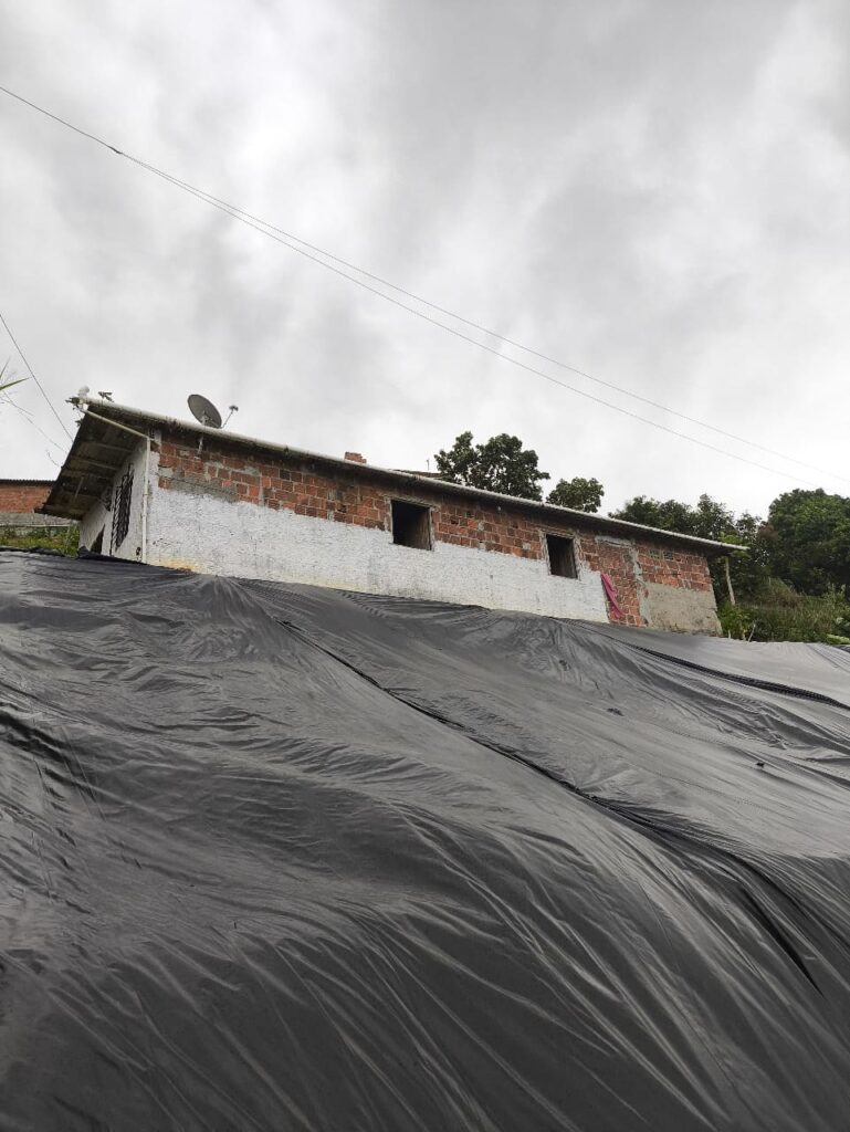 A imagem mostra uma casa simples de tijolos aparentes e paredes parcialmente pintadas de branco, localizada em uma área elevada. A casa tem um telhado inclinado com uma antena parabólica instalada no canto esquerdo. Há duas janelas sem vidros visíveis na parede frontal da casa. Em primeiro plano, há uma grande lona preta cobrindo o solo inclinado, possivelmente para proteger contra erosão ou deslizamentos de terra. O céu está nublado, e há algumas árvores ao fundo, indicando que a casa está em uma área rural ou periférica.