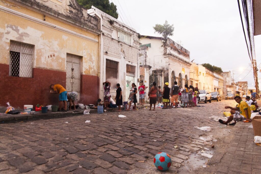 A imagem mostra uma rua de paralelepípedos em uma área urbana. À esquerda, há um prédio antigo com pintura desgastada e janelas gradeadas. No centro e à direita da imagem, há um grupo de pessoas, algumas em pé e outras sentadas, aparentemente conversando ou observando as atividades. No chão, há vários baldes, sacolas e objetos espalhados. Em primeiro plano, no canto inferior direito, há uma bola de futebol colorida.