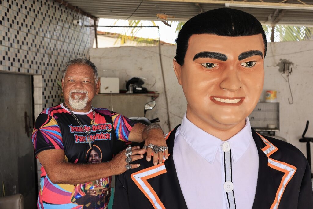 Foto de Kakay ao lado do boneco gigante Menino da Gráfica. Ele é um negro idoso, com cabelhos grisalhos, cavanhaque e bigode brancos, vestindo uma camisa colorida com a foto de uma mulher negra sobre a qual se lê em letras vermelhas Zenaide Bezerra - para eternizar. As mãos do homem, pousadas sobre o ombro do boneco, têm anéis prateados em todos os dedos. A foto foi feita durante o dia em um ambiente fechado cobertos com telhas de amianto, parecendo uma garagem ou galpão.