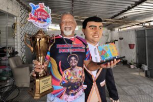 A imagem mostra um homem idoso negro, com barba, bigode e cabelo brancos, ao lado de um boneco gigante do carnaval de Olinda em um ambiente coberto, possivelmente uma garagem ou galpão. Ele está segurando com a mão direita um grande troféu dourado com uma placa que diz Campeão Geral 2023. O troféu tem um enfeite no topo com a inscrição Carnaval de Rua. Com a mão esquerda, ele segura uma placa com várias figuras coloridas. Ele está vestindo uma camiseta colorida com uma imagem de uma pessoa.